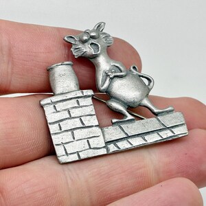 May include: A silver-toned metal brooch featuring a cartoon cat perched atop a brick chimney. The cat is mid-yawn, with its mouth open. The brooch has a pin on the back for attachment.