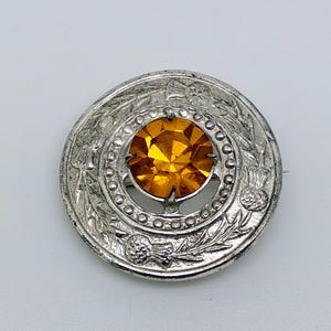 Puede incluir: Broche plateado con una piedra central facetada de color ámbar. El broche circular presenta un diseño ornamentado con motivos florales y de hojas alrededor del perímetro. Accesorio vintage.