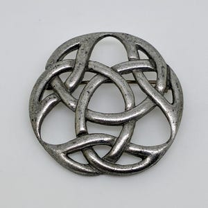 May include: A silver-toned Celtic knot brooch. The brooch features an intricate interwoven design, with a circular shape. The metal has a slightly aged appearance. The pin is a classic accessory.
