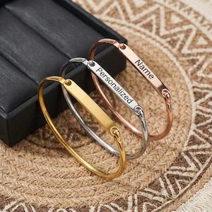 May include: Three personalized bangles in gold, silver, and rose gold. Each bangle features a rectangular plate with the words "Name" or "Personalized". The bangles are displayed on a textured surface.