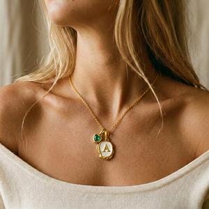 Op de afbeelding: Een goudkleurige ketting met een hanger met de letter "A" in wit emaille, omlijst door een getextureerde rand. Een kleine groene edelsteen hangt naast de hanger. De ketting wordt om de nek van een persoon gedragen.