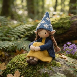 May include: A handmade felt doll of a seated gnome with a blue hat decorated with white snowflakes. The gnome wears a yellow dress and a blue jacket. The doll has brown braids and is sitting on a moss-covered rock in a forest setting.