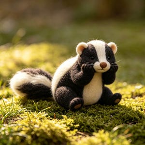 Op de afbeelding: Handgemaakte vilten stinkdierfiguur met een zwart-wit gestreept lichaam, zittend op groen mos. Het stinkdier heeft een witte streep op zijn hoofd en een bruine neus, met zijn poten bij zijn gezicht.