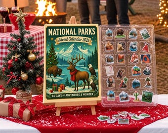 May include: A National Parks Advent Calendar 2026 box on a wooden easel, featuring a winter landscape with a deer, bison, and eagles. Beside it, a clear plastic tray holds twenty-four small, colorful nature-themed stickers and ornaments.