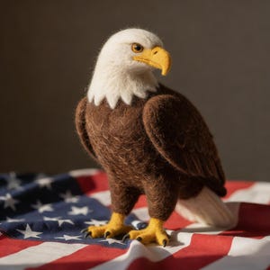 May include: A handcrafted felt bald eagle figurine with a white head, brown body, and yellow beak and feet. The eagle stands on a draped American flag, showcasing red and white stripes and blue field with white stars. The eagle is a symbol of American patriotism.