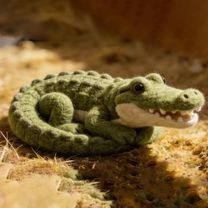 Puede incluir: Figura de cocodrilo de fieltro verde hecha a mano, con cuerpo texturizado y boca abierta que muestra dientes blancos. El cocodrilo está enroscado sobre hierba seca y musgo. Un elemento decorativo.