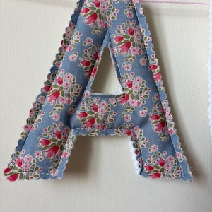 May include: A fabric letter A featuring a blue background with a repeating floral pattern of pink and red flowers with green leaves. The letter has scalloped edges with visible stitching and is suspended from a thin pink string against a wall.