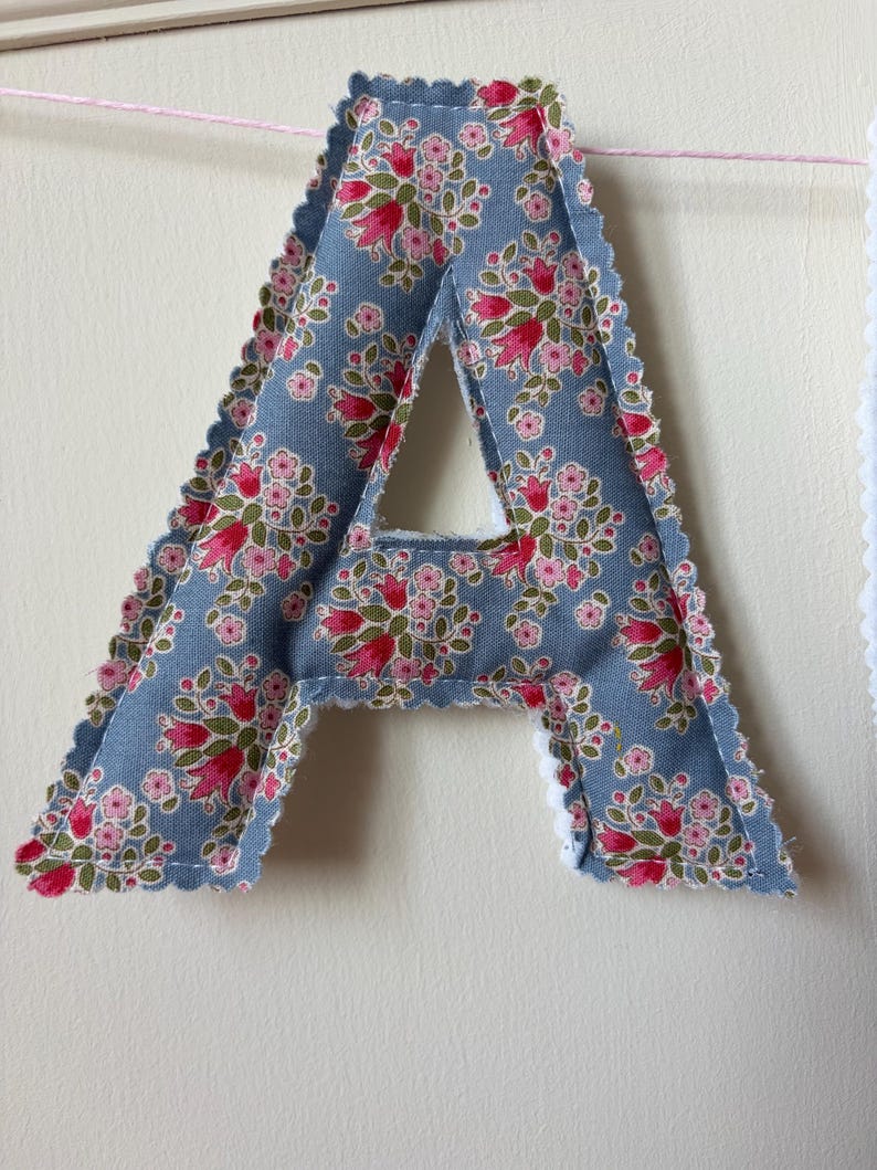 May include: A fabric letter A featuring a blue background with a repeating floral pattern of pink and red flowers with green leaves. The letter has scalloped edges with visible stitching and is suspended from a thin pink string against a wall.