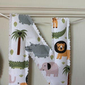 May include: A fabric letter N featuring a white background with a repeating pattern of colourful jungle animals including a lion, elephant, hippopotamus, and crocodile, along with palm trees and cacti, finished with a scalloped edge and white ribbon.