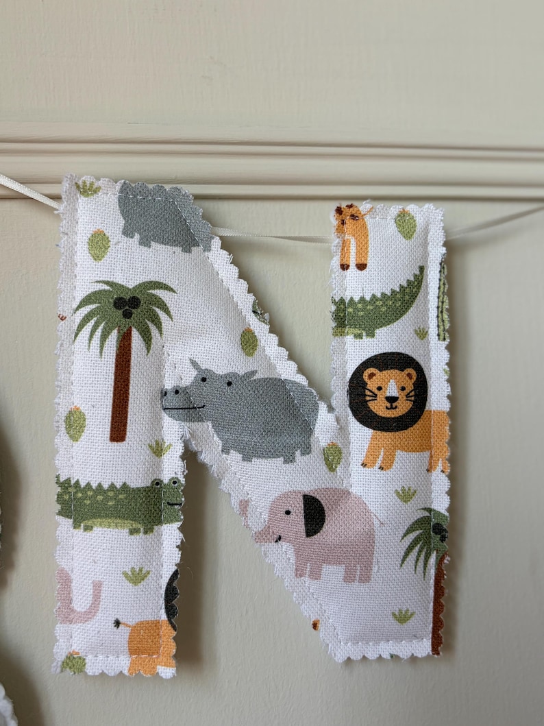May include: A fabric letter N featuring a white background with a repeating pattern of colourful jungle animals including a lion, elephant, hippopotamus, and crocodile, along with palm trees and cacti, finished with a scalloped edge and white ribbon.