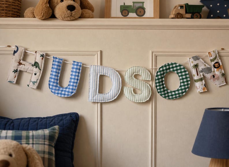 May include: A fabric banner spelling the name HUDSON, featuring individual stuffed letters in various patterns including dinosaur prints, blue and white gingham, light blue stripes, light green stripes, and dark green gingham, all strung on a white cord.