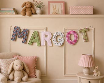 May include: A fabric banner spelling the name MARGOT in individual letters, each featuring a unique pattern including blue floral, green floral, pink striped, white floral, and pink gingham, suspended on a white string against a paneled wall.