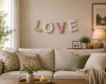 May include: A fabric banner spelling the word LOVE, featuring individual stuffed letters hanging on a string. The L is light green with a floral pattern, the O is white with small dark flowers, the V is pink gingham, and the E is white striped.