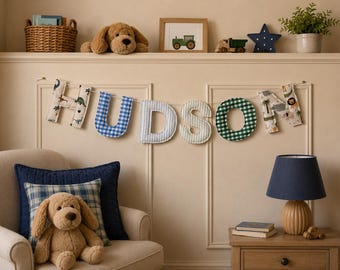 May include: A fabric letter banner spelling the name HUDSON, featuring individual letters in various patterns including dinosaur prints, blue and white gingham, light blue stripes, and green and white gingham, all strung together on a white cord.