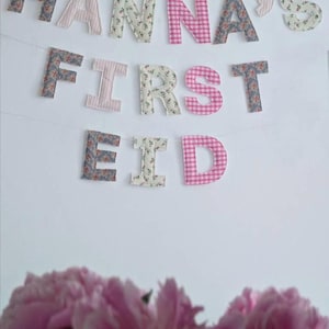 May include: A fabric banner featuring individual letters spelling out HANNA'S FIRST EID. The letters are crafted from various patterned fabrics including floral prints, pink and white stripes, and pink and white gingham checks, all strung together.