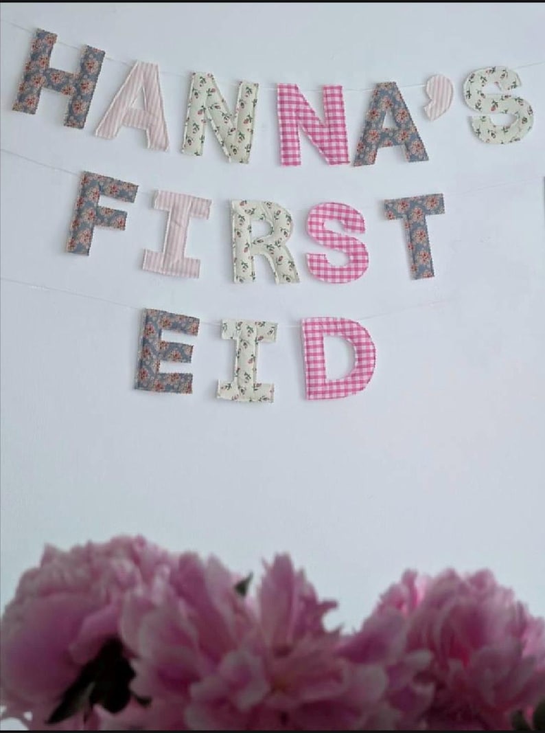 May include: A fabric banner featuring individual letters spelling out HANNA'S FIRST EID. The letters are crafted from various patterned fabrics including floral prints, pink and white stripes, and pink and white gingham checks, all strung together.