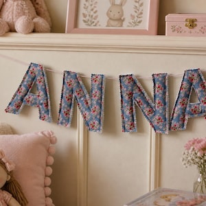May include: A fabric banner spelling the name ANNA, featuring individual stuffed letters made from blue fabric with a red and white floral pattern, finished with white scalloped edges and strung together on a thin white cord.