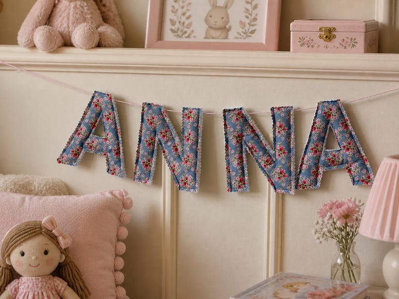 May include: A fabric banner spelling the name ANNA, featuring individual stuffed letters made from blue fabric with a red and white floral pattern, finished with white scalloped edges and strung together on a thin white cord.
