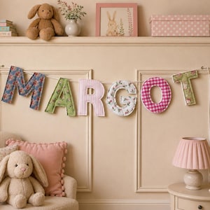 May include: A fabric banner spelling the name MARGOT in individual letters, each featuring a unique pattern including blue floral, green floral, pink striped, white floral, and pink gingham, suspended on a white string against a paneled wall.