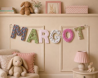May include: A fabric letter banner spelling the name MARGOT, featuring individual letters in various floral, gingham, and solid pastel patterns. Each letter is padded and suspended on a white string, creating a decorative wall hanging display.