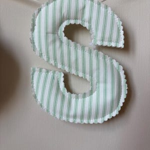 May include: A stuffed fabric letter S featuring a light green and white vertical striped pattern. The letter has a scalloped edge with visible stitching and is suspended by a thin white ribbon, creating a soft, padded appearance for display.