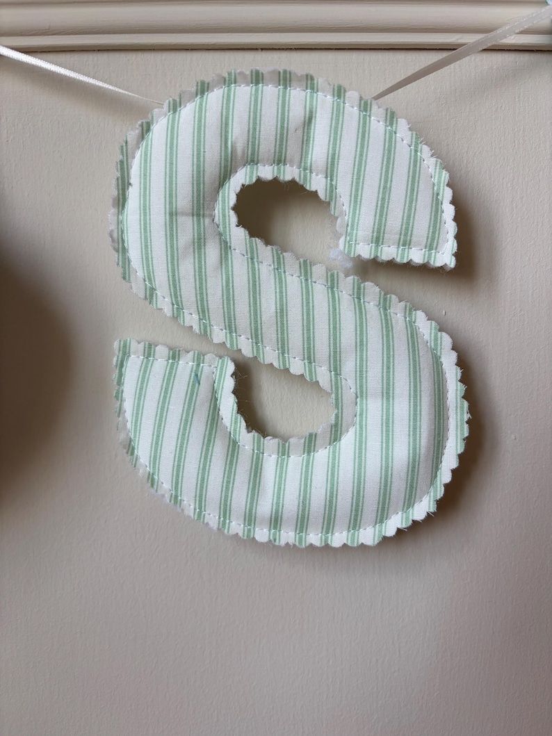 May include: A stuffed fabric letter S featuring a light green and white vertical striped pattern. The letter has a scalloped edge with visible stitching and is suspended by a thin white ribbon, creating a soft, padded appearance for display.