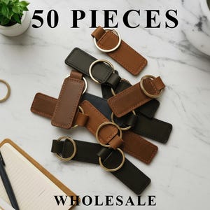 May include: An assortment of leather keychains in brown, black, and green, each with a gold-colored ring. The text "50 PIECES WHOLESALE" is displayed in large, bold letters above and below the keychains.
