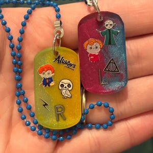 Puede incluir: Dos colgantes rectangulares de resina en una cadena de bolas azul. El colgante amarillo presenta un personaje de dibujos animados, un búho, un rayo, la letra R y el texto Always. El colgante rosa y azul presenta dos personajes y un símbolo.