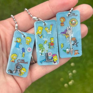 Può includere: Tre portachiavi rettangolari in resina azzurra con personaggi e icone dei Simpson. Ogni pezzo include una catenella a palline in metallo, lettere R, L e una freccia in metallo incastonate, oltre a vari piccoli adesivi di personaggi.