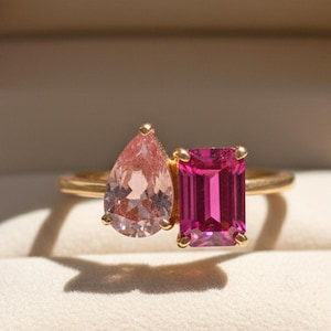 Pode incluir: Um anel de ouro com duas pedras preciosas: uma morganita rosa em forma de pêra e uma turmalina rosa com corte esmeralda. O anel é apresentado dentro de uma caixa de joias castanha com interior creme.