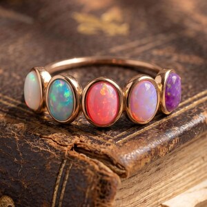 Puede incluir: Un anillo de tono dorado con cinco piedras cabujón de forma ovalada dispuestas en fila. Las piedras muestran una variedad de colores que incluyen blanco, azul verdoso iridiscente, rojo anaranjado vibrante, rosa suave y morado intenso, montadas en una banda metálica fina.