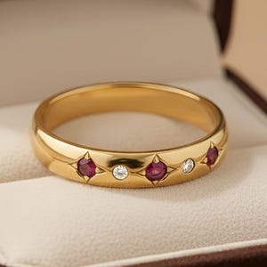 Puede incluir: Un anillo de oro con tres piedras rojas y dos piedras transparentes, presentado en un joyero marrón. El anillo es el punto focal, con el texto "NOVA JEWELS" visible en el interior.