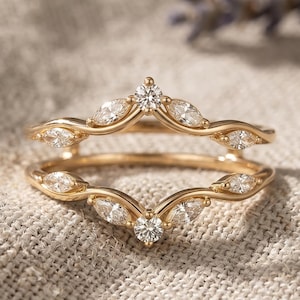 May include: A gold ring guard featuring two curved bands that meet at a central point. The bands are set with marquise-cut and round-cut clear stones arranged in a symmetrical pattern along the top and bottom edges of the open design.