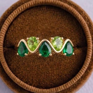May include: A gold ring featuring a series of green gemstones. The ring has a wavy design with three pear-shaped emeralds and two round, light green peridot stones. The ring is presented in a brown velvet ring box.