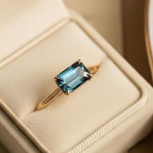 Puede incluir: Un anillo de oro con una gema azul rectangular se muestra en un joyero crema. La caja está abierta, revelando el engaste del anillo. La caja está sobre un papel vintage con escritura y un plato blanco con borde dorado.