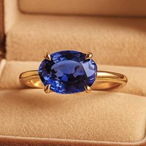 Op de afbeelding: Een gouden ring met een grote, ovale, diepblauwe edelsteen, gepresenteerd in een open bruine fluwelen doos. De doos staat op vintage papier met handschrift. De tekst "NOVA JEWELS" is in de doos geëmbosseerd. Een sieraad.