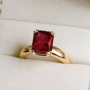 Puede incluir: Un anillo de oro con una gran gema roja oscura rectangular. El anillo se presenta en un joyero de terciopelo blanco abierto. La gema es el punto focal, con un estilo clásico.