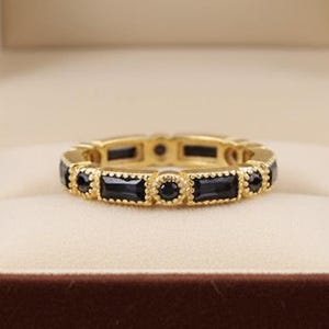 Pode incluir: Um anel dourado com pedras pretas retangulares e redondas alternadas. O anel é apresentado numa caixa de joias de veludo castanho com interior creme.