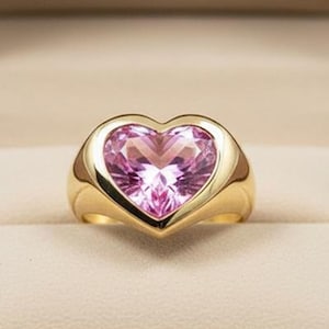 Könnte beinhalten: Ein goldener Ring mit einem großen, herzförmigen rosa Edelstein. Der Ring wird in einer offenen braunen Schmuckschatulle mit dem Text "NOVA JEWELS" auf der Innenseite des Deckels präsentiert. Der Ring hat ein dickes, poliertes Band.