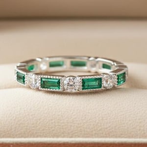 Puede incluir: Un anillo plateado con piedras preciosas verdes de talla esmeralda y piedras blancas redondas. El anillo se muestra en una caja de joyería marrón con el texto "NOVA JEWELS" en letras doradas.