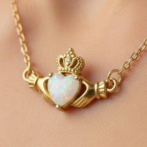 May include: A gold Claddagh necklace featuring a heart-shaped opal stone, a crown, and hands. The necklace hangs on a fine chain. The heart is white with a shimmering effect. The crown is detailed and sits above the heart.