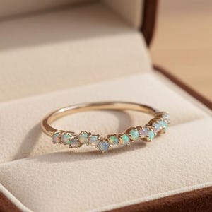 May include: A gold ring with a row of small opals set in a curved design, displayed inside a brown jewelry box with a cream-colored interior. The ring is a delicate piece of jewelry.