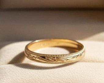May include: A gold wedding band with an engraved floral pattern, displayed in a brown velvet ring box. The ring is centered on a cream-colored cushion, with the box open on a wooden surface. The ring's design includes a series of small dots.