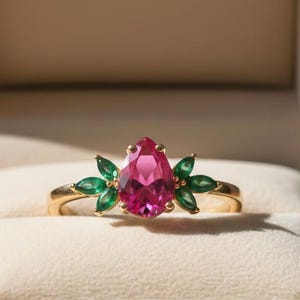 May include: A gold ring with a pear-shaped pink gemstone as the centerpiece, flanked by green gemstones. The ring is presented in a brown velvet jewelry box with a cream-colored interior. The ring's design is floral-inspired.