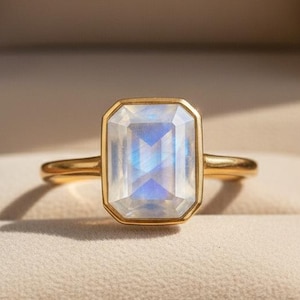 May include: A gold ring featuring a rectangular, iridescent gemstone, set in a gold bezel. The ring is presented in a brown jewellery box with the text "NOVA JEWELS" embossed on the inside of the lid.