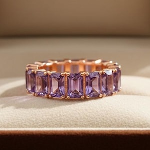以下が含まれることがあります： 茶色のベルベットのジュエリーボックスに入った、長方形の紫色の宝石が付いたローズゴールドのリング。 ボックスの蓋が開いており、クリーム色のクッションの上にリングが置かれています。 ボックス内には「NOVA JEWELS」の文字が見えます。