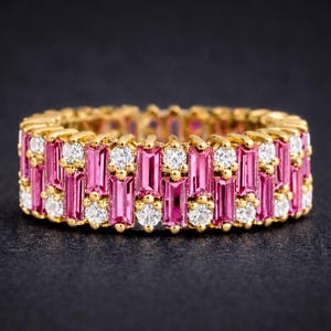 Könnte beinhalten: Ein goldfarbener Ring mit abwechselnden Reihen aus rosa Baguette-Edelsteinen und runden, klaren Edelsteinen. Der Ring ist vor einem dunklen Hintergrund platziert, der das Funkeln der Steine hervorhebt. Der Ring ist ein Schmuckstück.