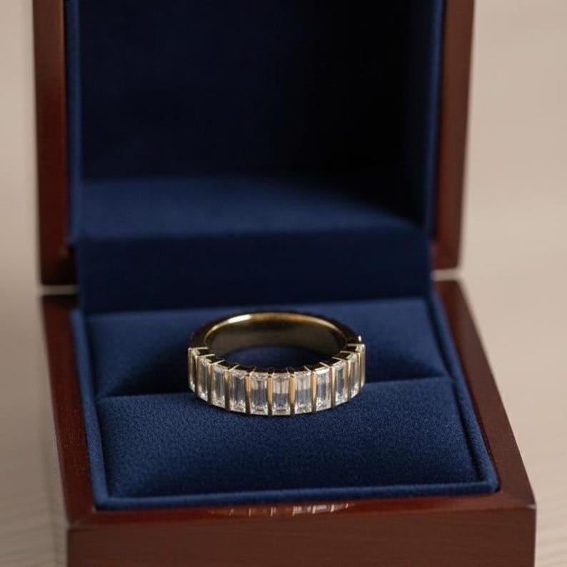 May include: A gold ring with rectangular, clear stones set around the band. The ring is displayed inside a dark blue velvet-lined jewelry box with a wooden exterior.