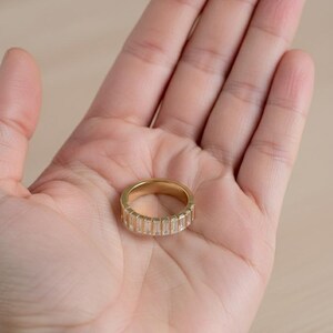 May include: A gold-colored ring with a band of rectangular, clear stones. The ring is held in an open palm, showcasing its design. The ring's band is approximately 0.25 inches wide. The ring is a piece of jewelry.