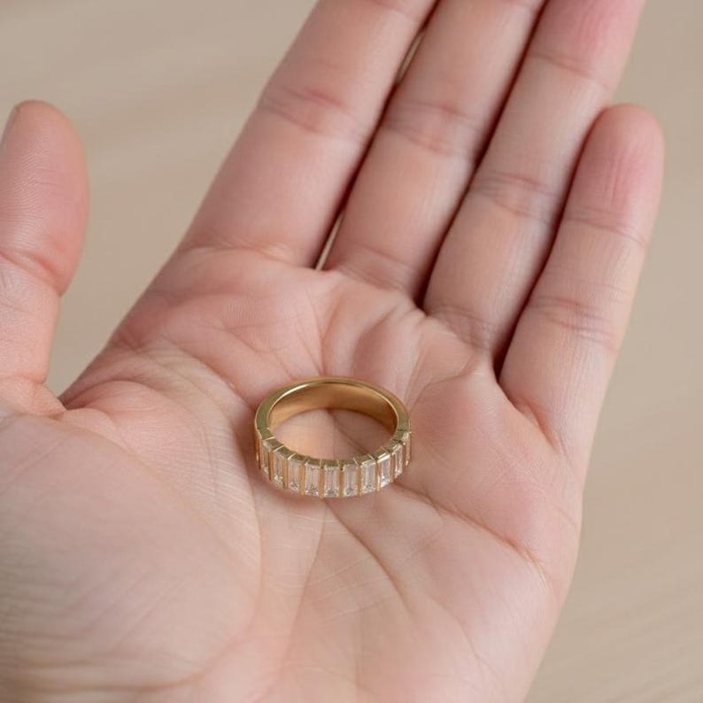 May include: A gold-colored ring with a band of rectangular, clear stones. The ring is held in an open palm, showcasing its design. The ring's band is approximately 0.25 inches wide. The ring is a piece of jewelry.